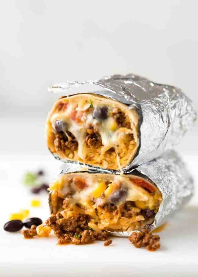 Beef burritos wrapped in foil cut in half with beef, rice, veggies, and sauce filling.