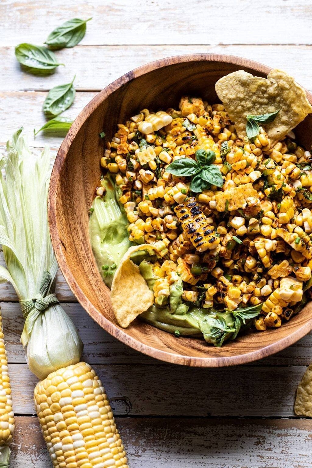 Grilled corn salad on a wooden bowl with chips, avocado dressing and basil leaves.