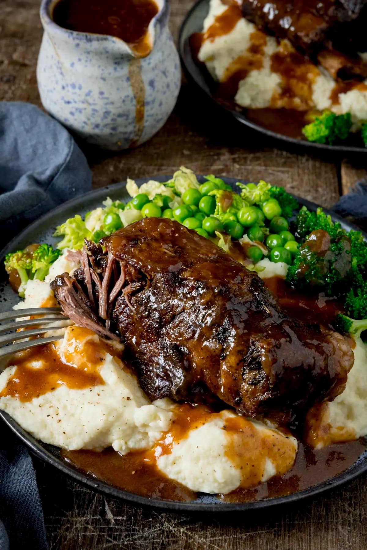Mashed potatoes topped with short ribs and gravy with green peas, broccoli and cauliflower on side.